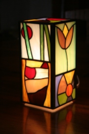 Lampe "Naïve" contemporaine en vitrail Tiffany. Ses représentations figuratives naïves confèrent à cette lampe une allure très gaie, lumineuse et très colorée. Elle est constituée de verres opalescents multicouleurs.