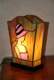 Lampe "L'Elegante" en vitrail Tiffany. Verre opalescent de couleur rose/blanc et chair pour le personnage et vert, bleu, rose et jaune doré pour le fond. Visage peint à la grisaille et cuit à 600°. Largeur bas 16 cm Largeur haut 21 cm