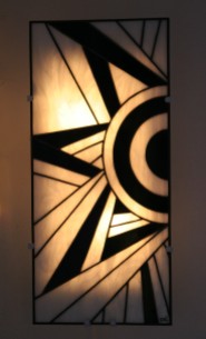 "Ycara" est une applique murale en vitrail Tiffany (hauteur 56 cm, largeur 26 cm). Son dessin s'inspirant de la géométrie cubiste donne à cette applique son modernisme mais aussi son style intemporel inspiré de l'Art Déco. Les verres, opalescents et de couleur Blanc nacré et Noir donneront à votre intérieur une ambiance à la fois lumineuse et chaleureuse. "Ycara" est constituée de 38 pièces de verre montées en vitrail Tiffany, et les soudures, à l'étain, ont été patinées en noir pour un aspect ancien. Elle est montée sur une structure électrifiée en acier blanc (fournie), équipée de 2 encoches de suspension pour un accrochage aisé sur un mur disposant d'une arrivée électrique.