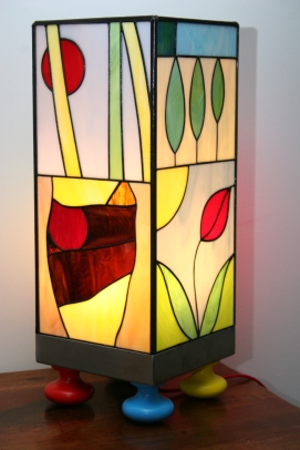 Originale et Unique : "Candide" est une très grande Lampe (52 cm) d'Art Naïf Contemporain en Vitrail Tiffany. Les verres sont opalescents et multicolores (bleus, rouges, oranges, noir, violets, jaunes, verts, marrons....). Très vive, gaie, colorée et lumineuse, cette lampe est constituée d'un socle en inox brossé monté sur 4 pieds de bois peint vert, rouge, bleu et jaune pour renforcer son côté ludique et joyeux. Les 4 faces sont toutes différentes et faites pour chacune d'entre elles de 2 scènes naïves stylisées et très colorées en véritable vitrail Tiffany.